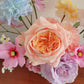 Assorted flowers including a peach-colored rose and pink and purple blooms on a wooden surface.