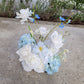 Arrangement of white and light blue flowers on a concrete surface with greenery in the background.