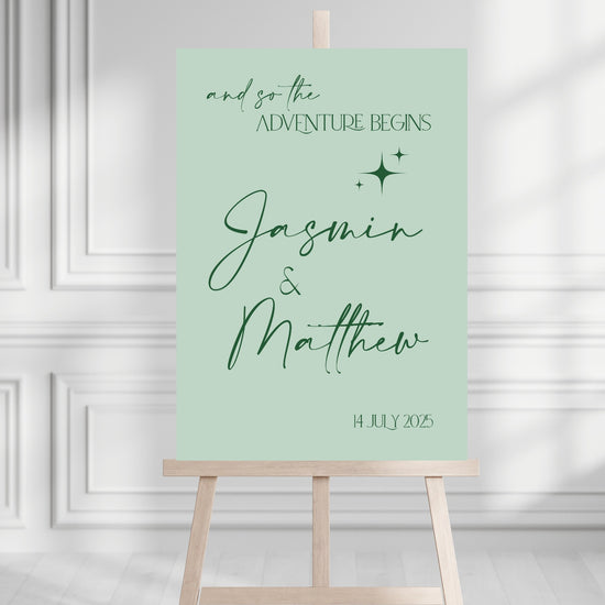 Green welcome sign with 'and so the adventure begins' text on a wooden stand against a white wall.