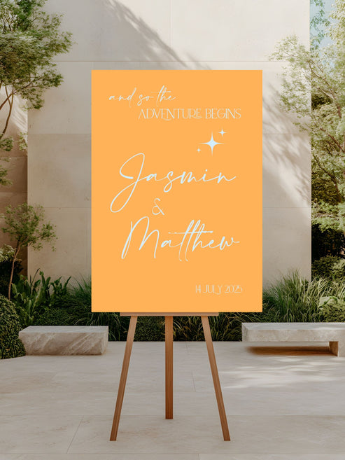 Orange sign with 'and so the adventure begins' and 'Jasmin & Matthew' on an easel outdoors.