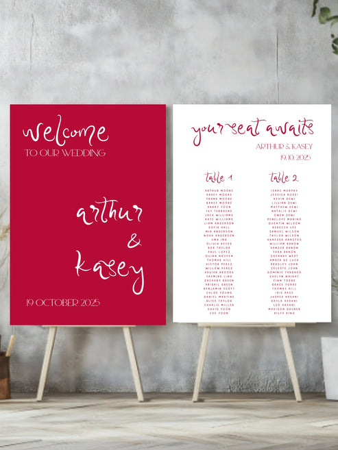Red welcome sign and white seating chart for a wedding on stands against a gray wall.