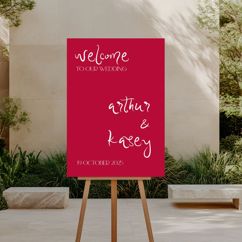 Wedding welcome sign on an easel outdoors with a red background and white text.