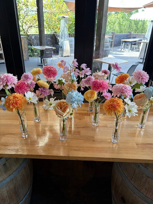 A collections of bud vases with colourful flowers on a wooden bench in front of a window