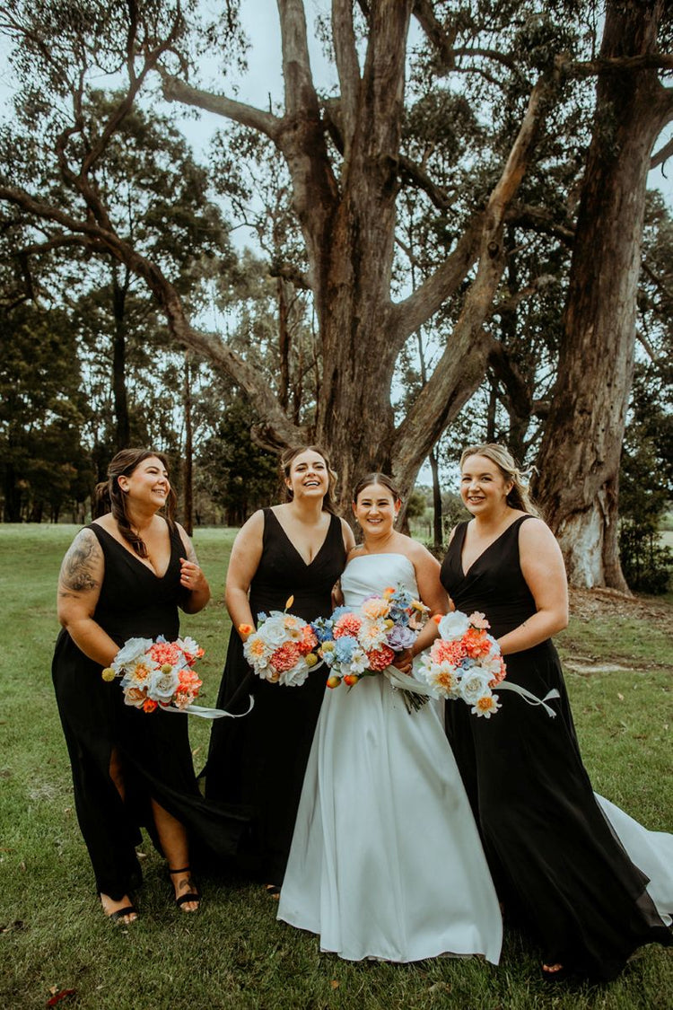 Bride with three bridesmaids in black dresses standing in a grassy area with trees.