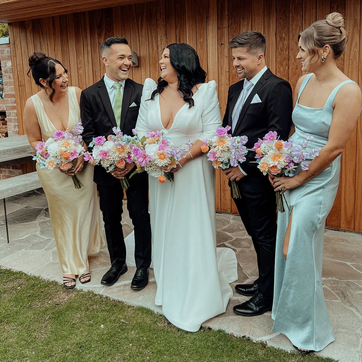 Group of people in formal attire with floral bouquets standing in front of a wooden building.