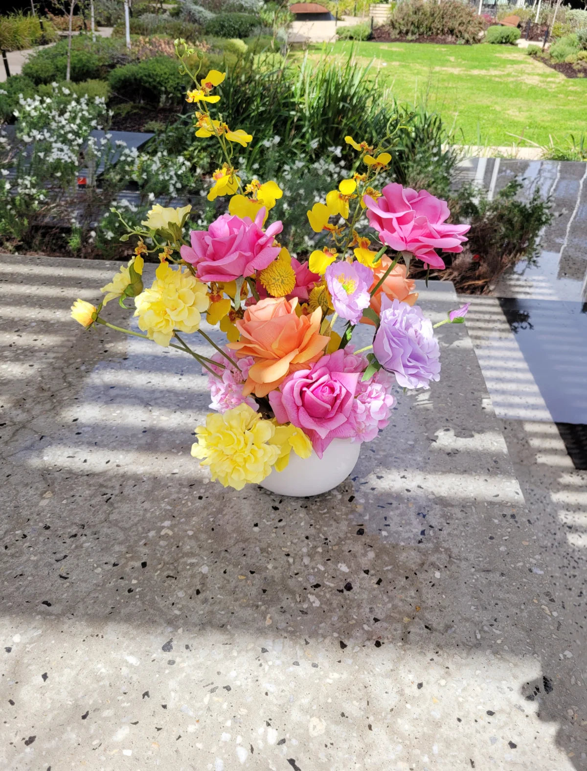 Colourful flower arrangement in a pot on a concrete surface with greenery in the background