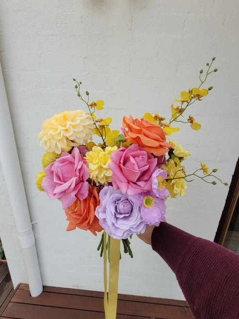 Bouquet of colorful artificial flowers held by a person against a white wall.