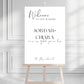 Wedding welcome sign on a wooden stand against a gray wall.