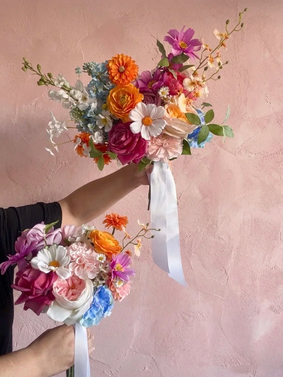 Two colourful artificial wedding bouquets held up by hands against a pink textured wall