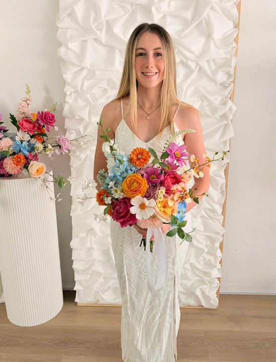 Woman in a white dress holding a colorful bouquet of artificial flowers against a decorative white wall.