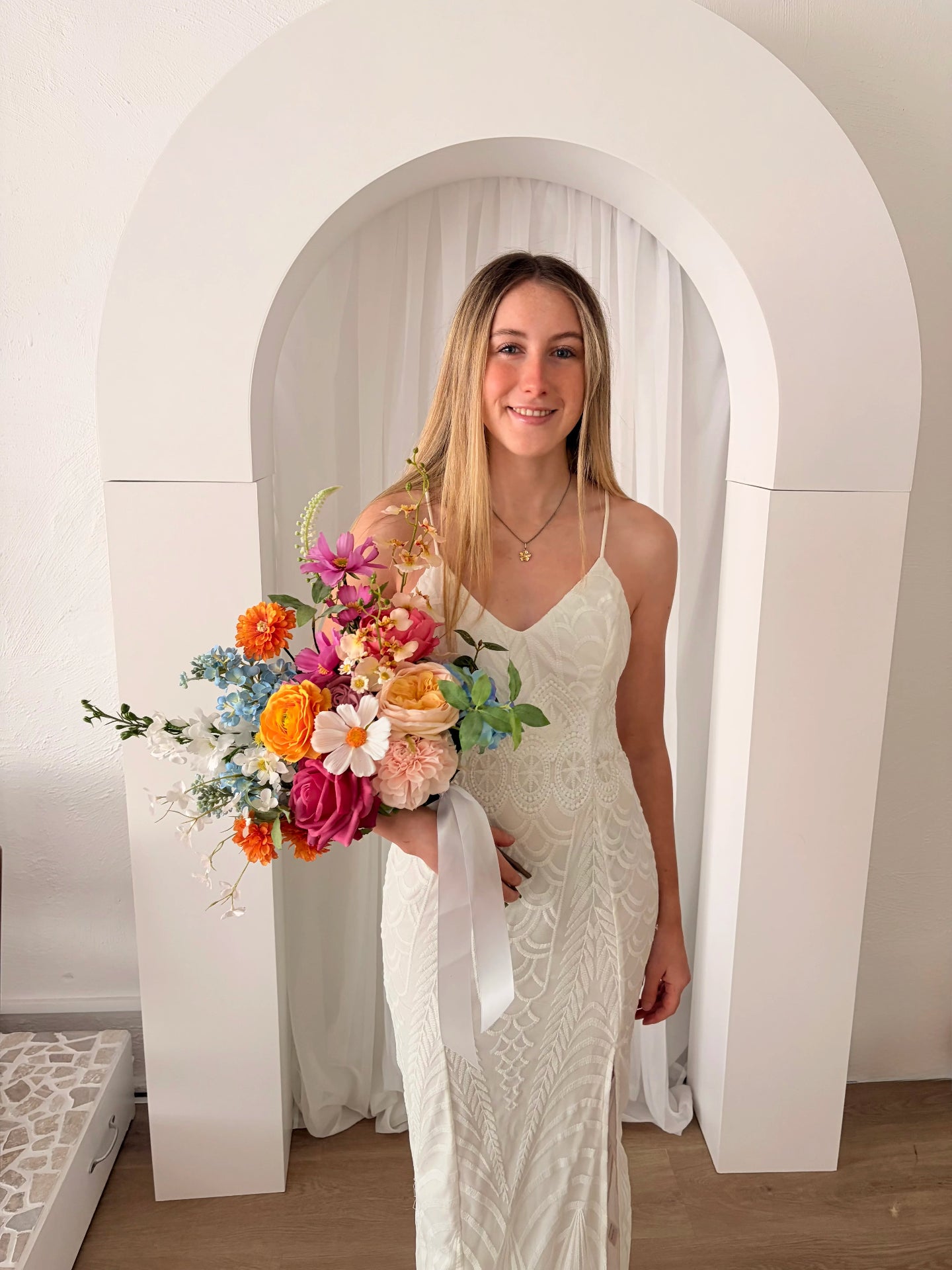 Woman in a white dress holding a colorful faux bouquet in front of a white archway.