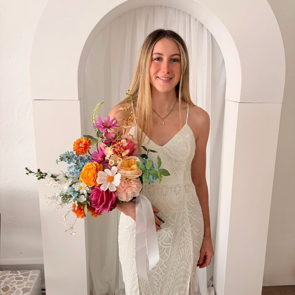Woman in a white dress holding a colourful wedding bouquet in front of a white archway.