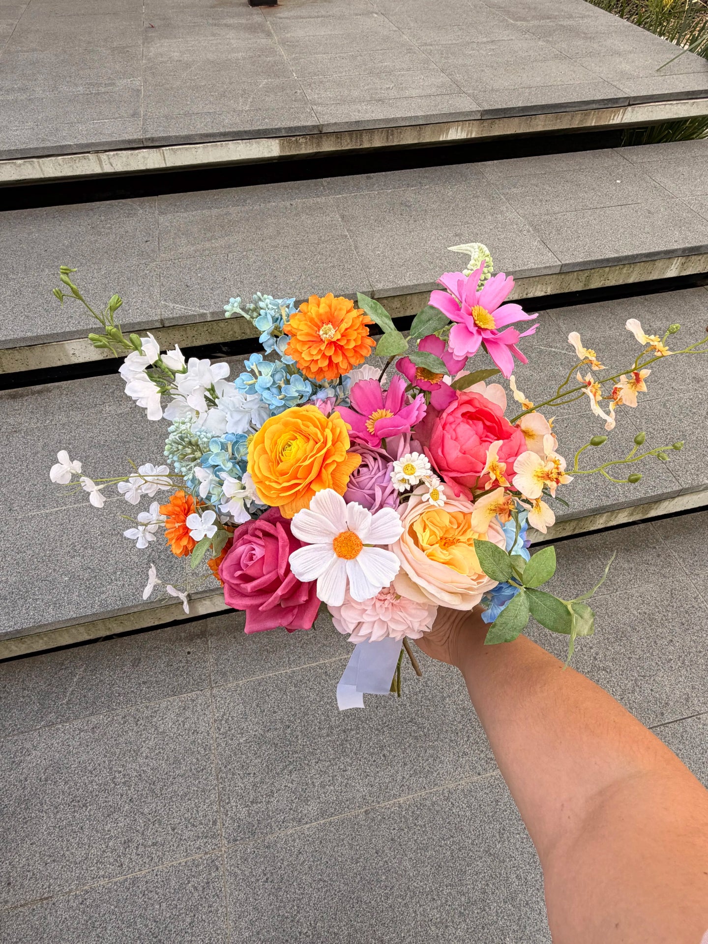Artificial wedding bouquet of colourful faux flowers held by a person on a stone step background