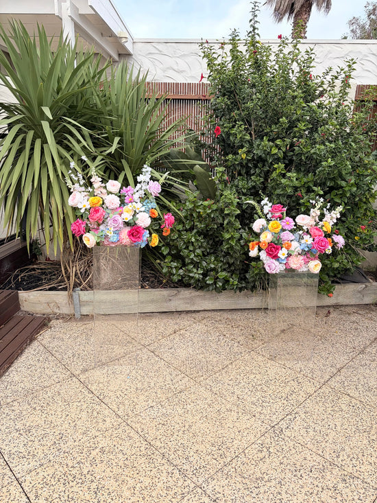 Colourful artificial flower arrangements on wedding plinths with plants and a building in the background.