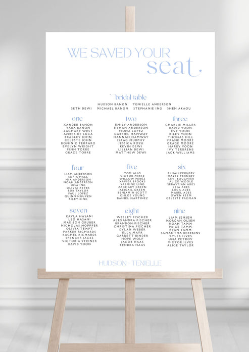 Seating chart for a wedding on a white board with a wooden stand against a gray background
