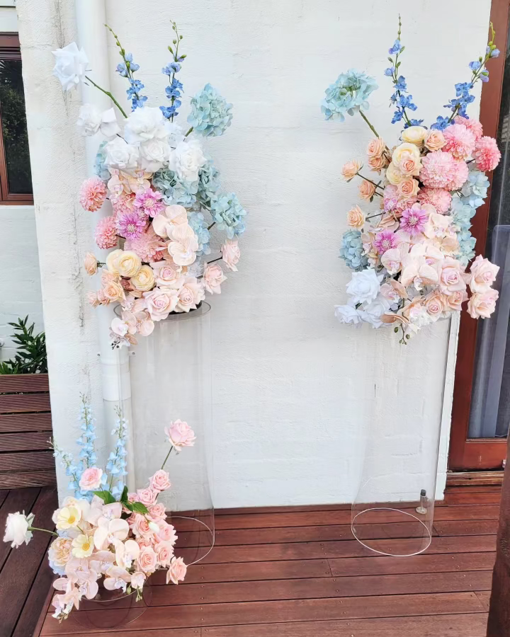 Decorative plinth floral arrangements on clear stands against a white brick wall.