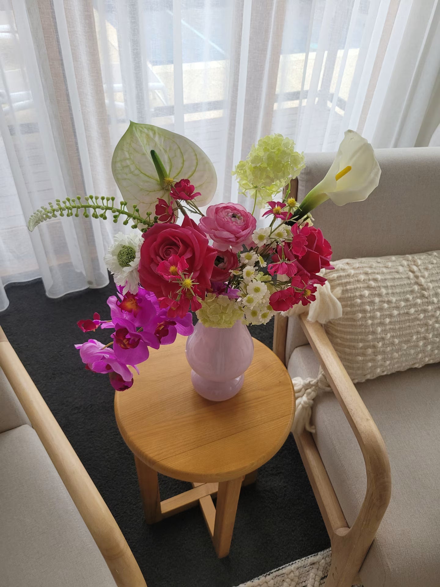 Floral arrangement in a pink vase on a wooden side table with a white sofa and curtains in the background.