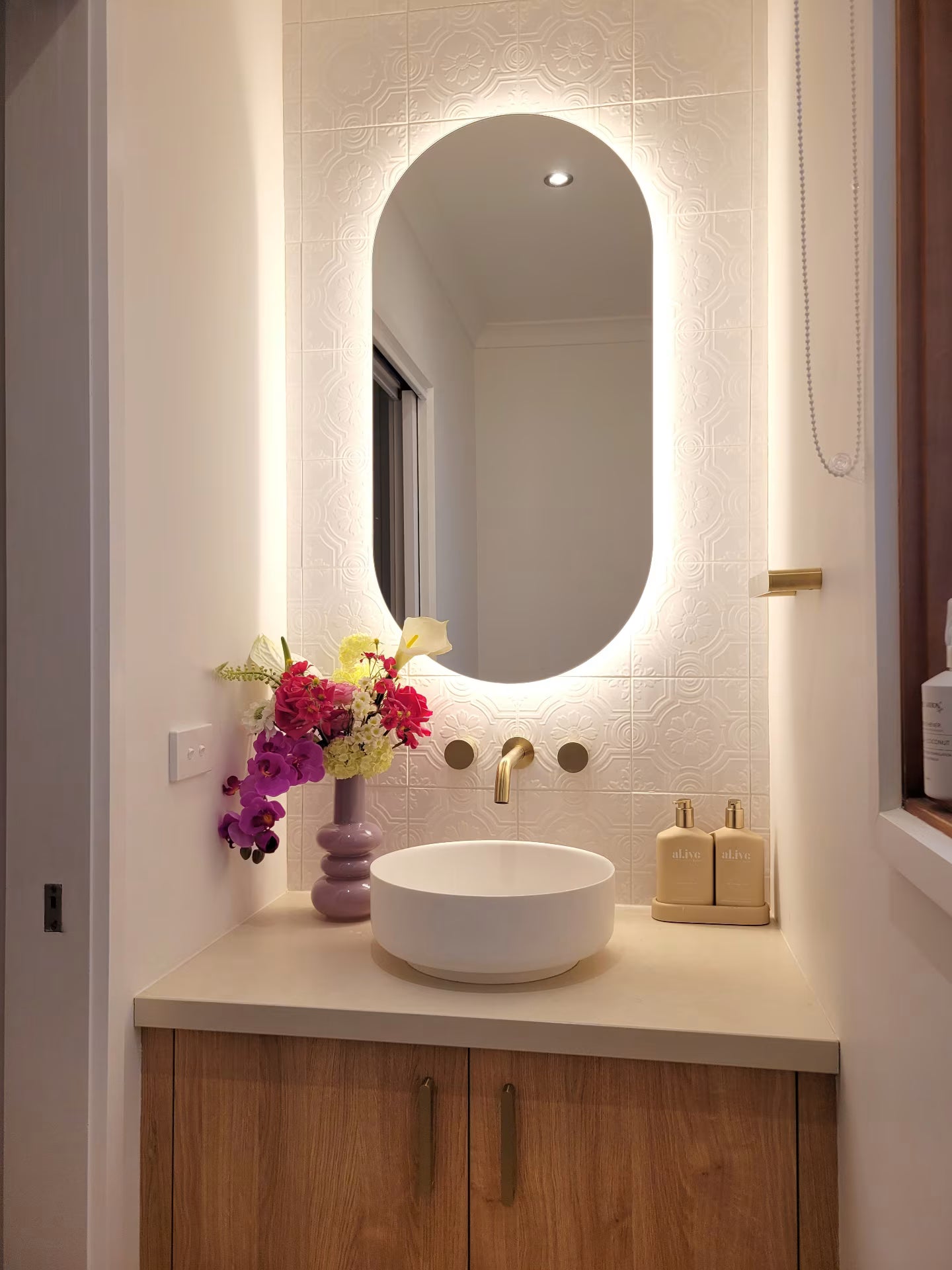 Bathroom vanity with illuminated mirror, sink, and a floral arrangement in the corner