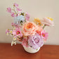 Bouquet of pink, orange, and purple flowers in a white vase on a wooden surface with a beige wall background.