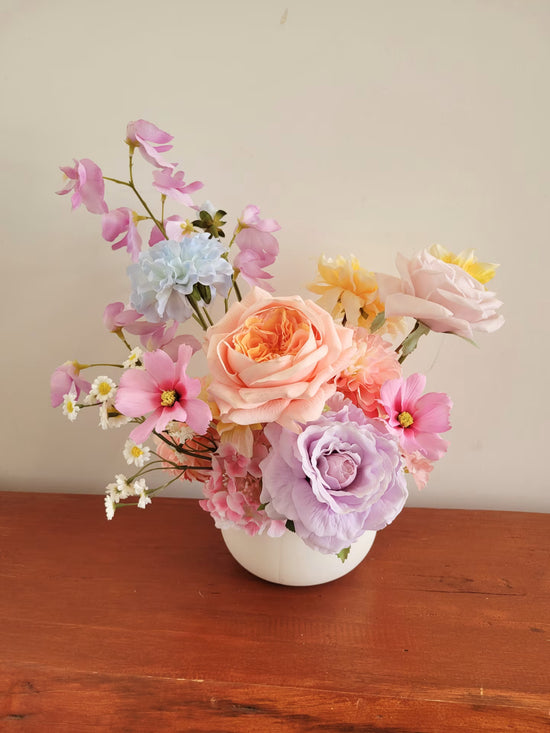 Bouquet of pink, orange, and purple flowers in a white vase on a wooden surface with a beige wall background.