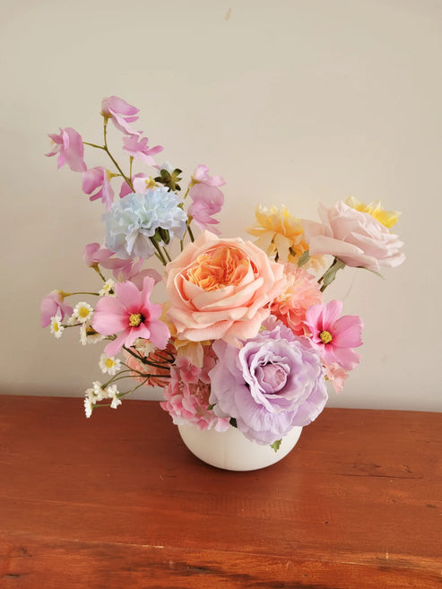 Assorted flowers in a white vase on a wooden surface with a neutral background
