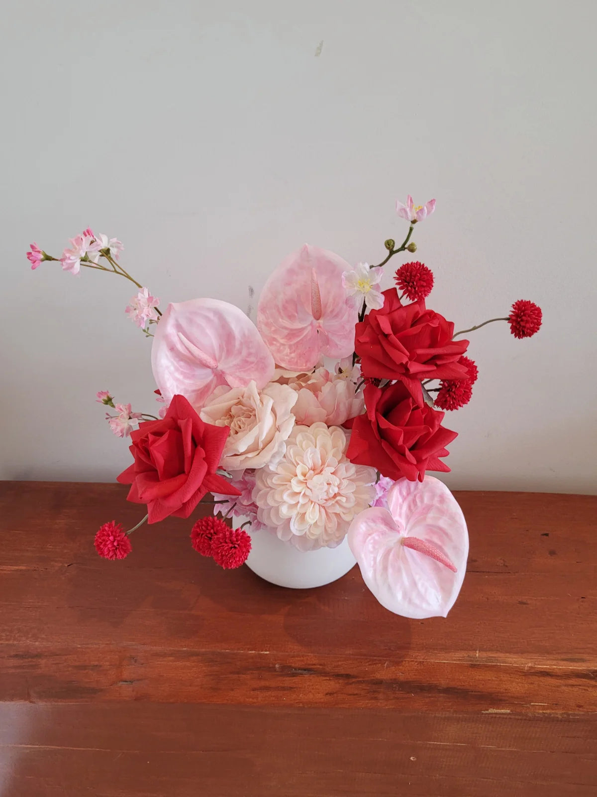 Faux flower arrangement of red and pink flowers in a white pot on a wooden surface