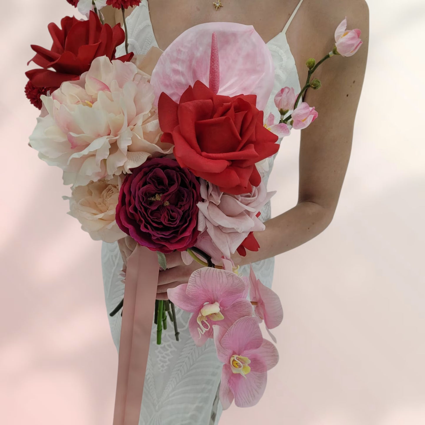 Bridal bouquet of red, pink, and white flowers held by a person against a soft pink background