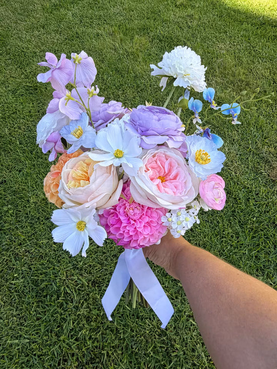 Artificial wedding bouquet of pastel flowers held by a hand over grass