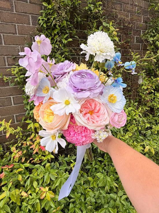 Bridal bouquet of pastel flowers held by a person against a brick wall and greenery background
