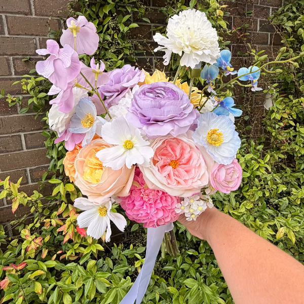Bouquet of colourful artificial flowers held by a person against a brick wall and greenery background
