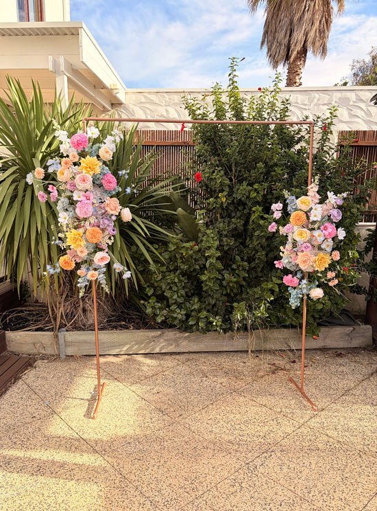 Two floral arrangements on a copper pipe arbour in a garden setting with plants and a building in the background.