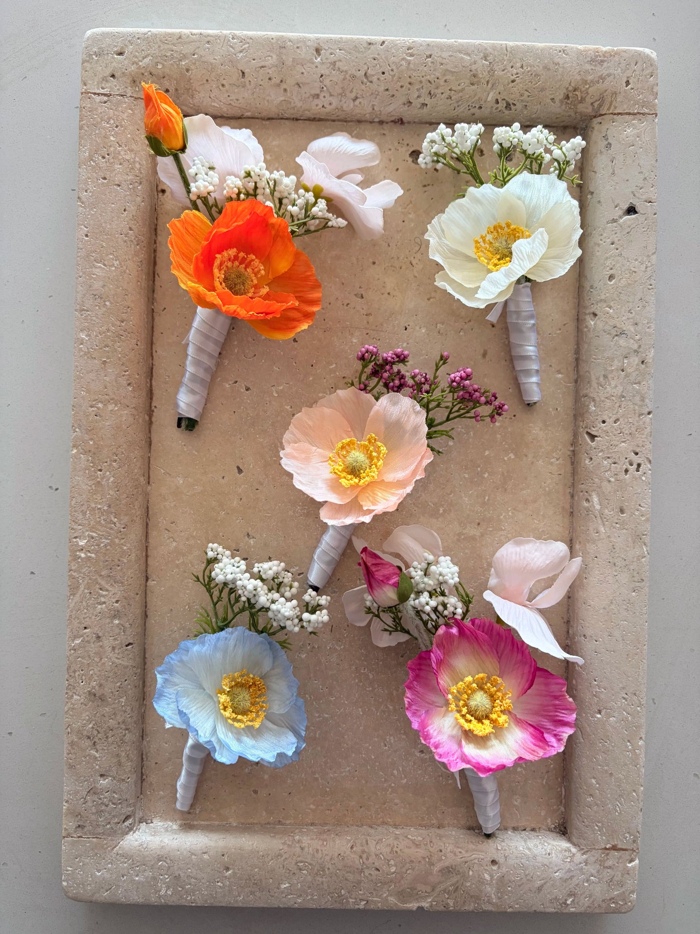 Five poppy buttonholes in orange, white, peach, blue and pink on a travertine display tray