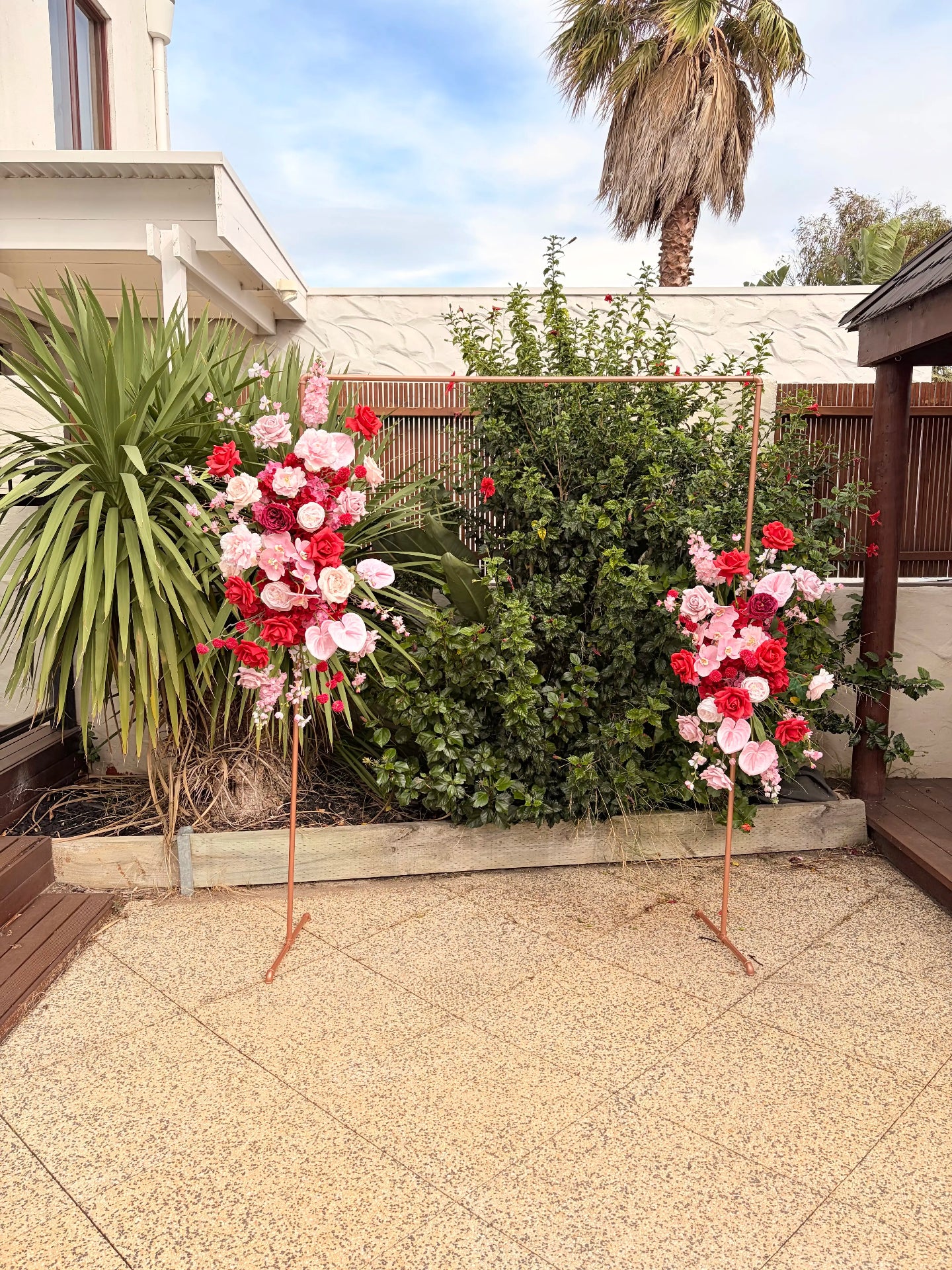 Two silk floral arrangements on stands in a garden setting with a white house and palm tree in the background.