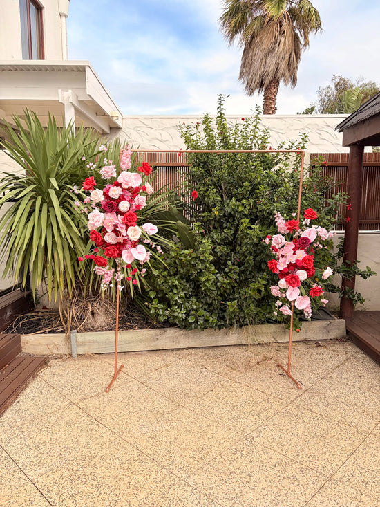 Two silk floral arrangements on stands in a garden setting with a white house and palm tree in the background.