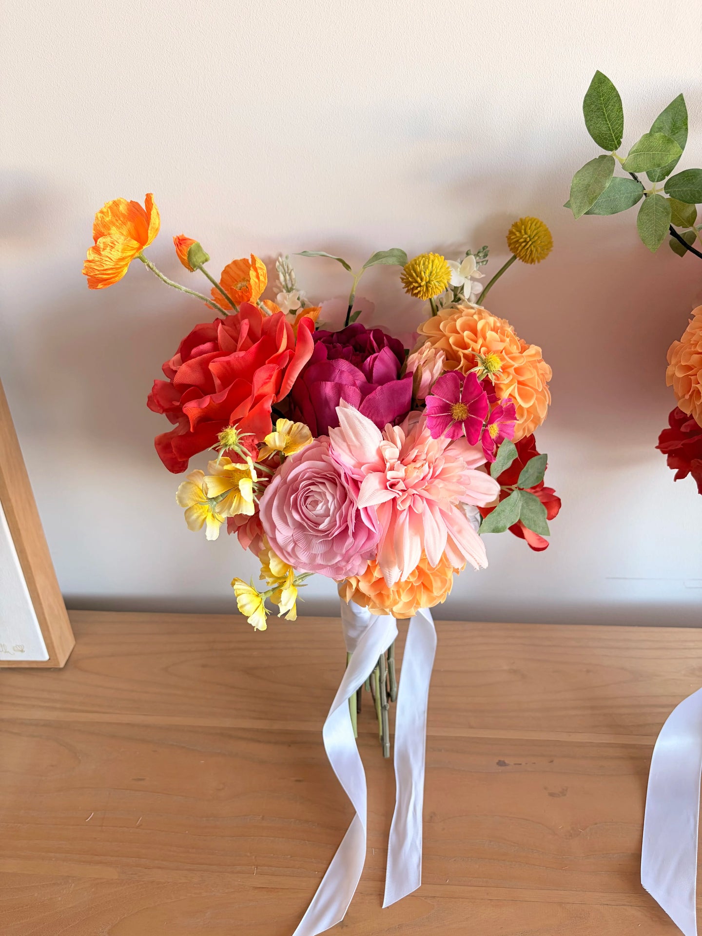 Artificial wedding bouquet for a bridesmaid in fuchsia, orange and pink leaning against a white wall.