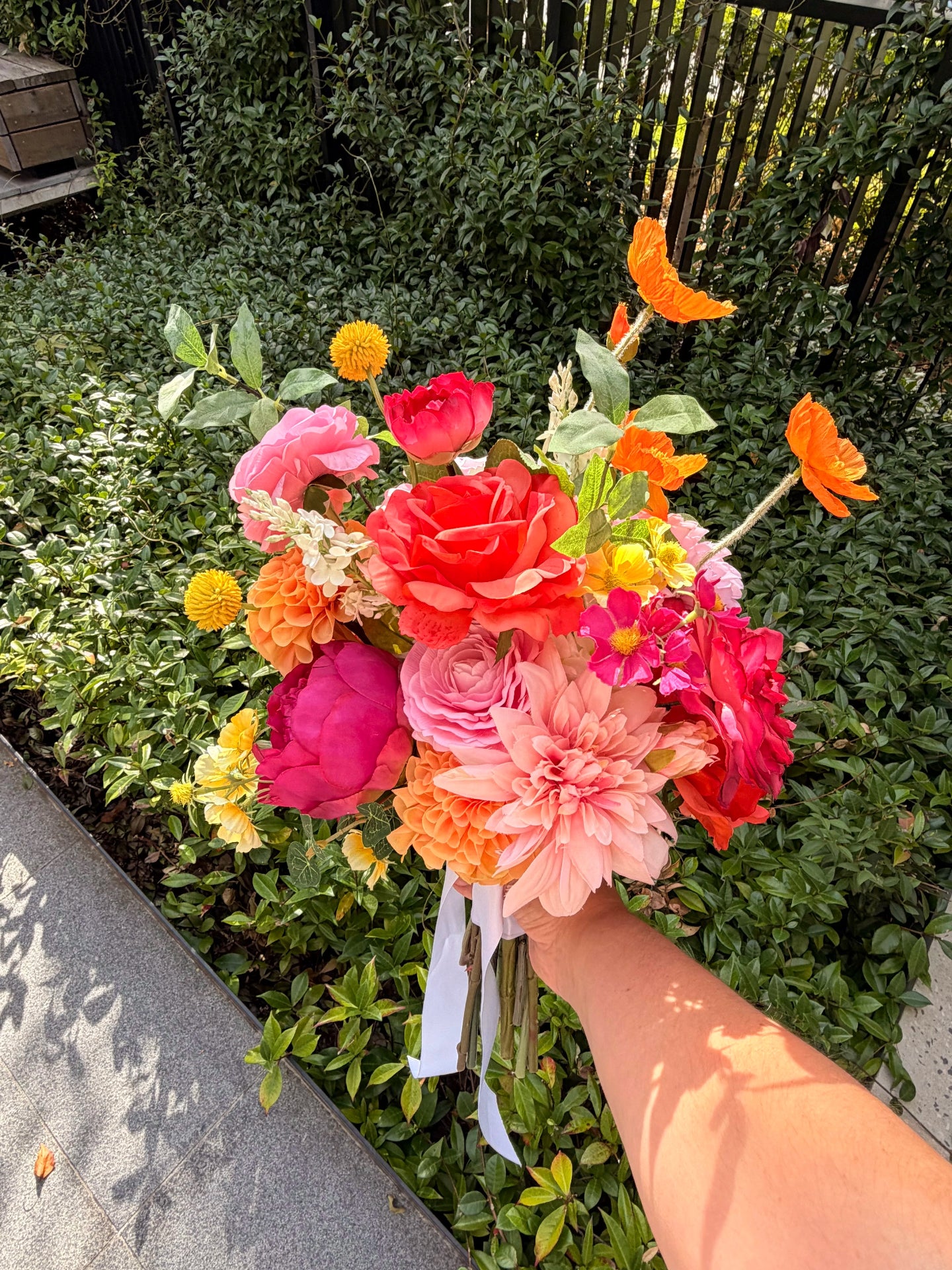 Faux wedding bouquet of colourful flowers held by a person in front of green foliage