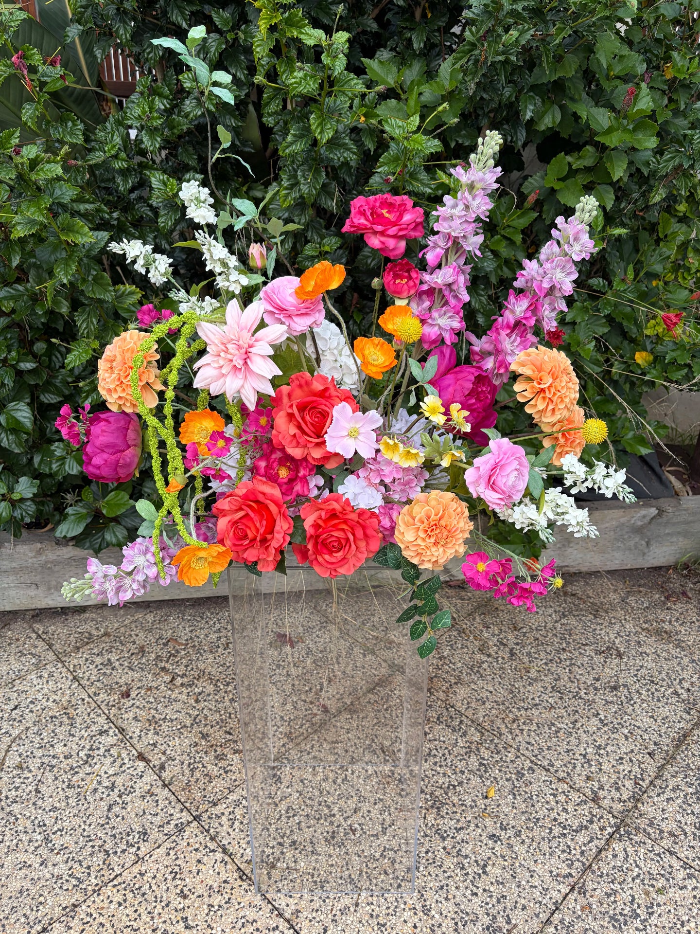 Colourful faux flower arrangement on a clear plinth on a stone surface with greenery in the background