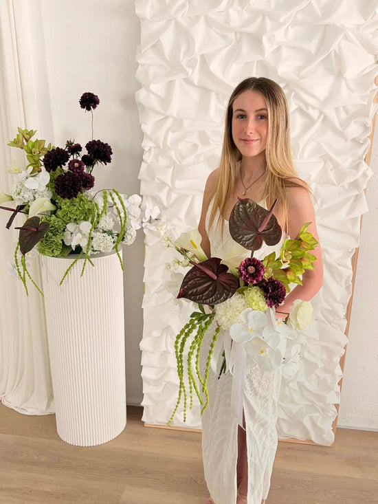 Woman holding a sculptural floral arrangement in front of a decorative white wall.