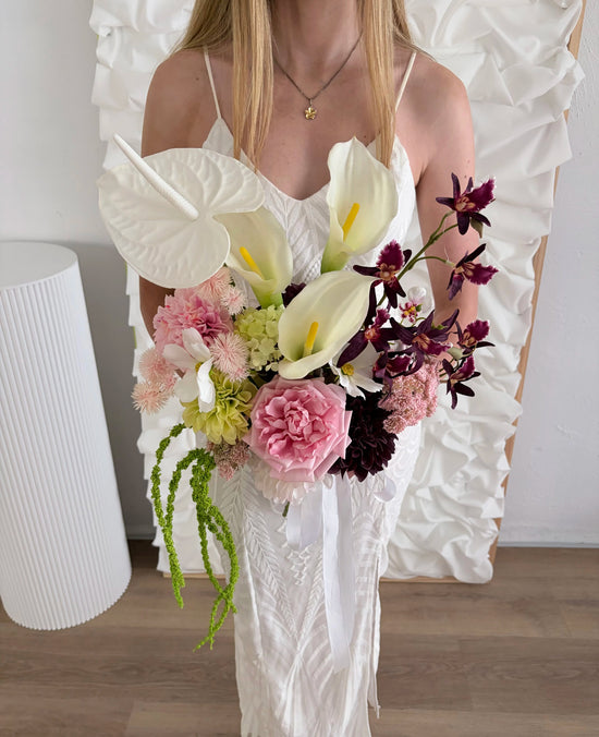 Woman holding a bouquet of flowers with prominent calla lilies in a white dress against a plain background