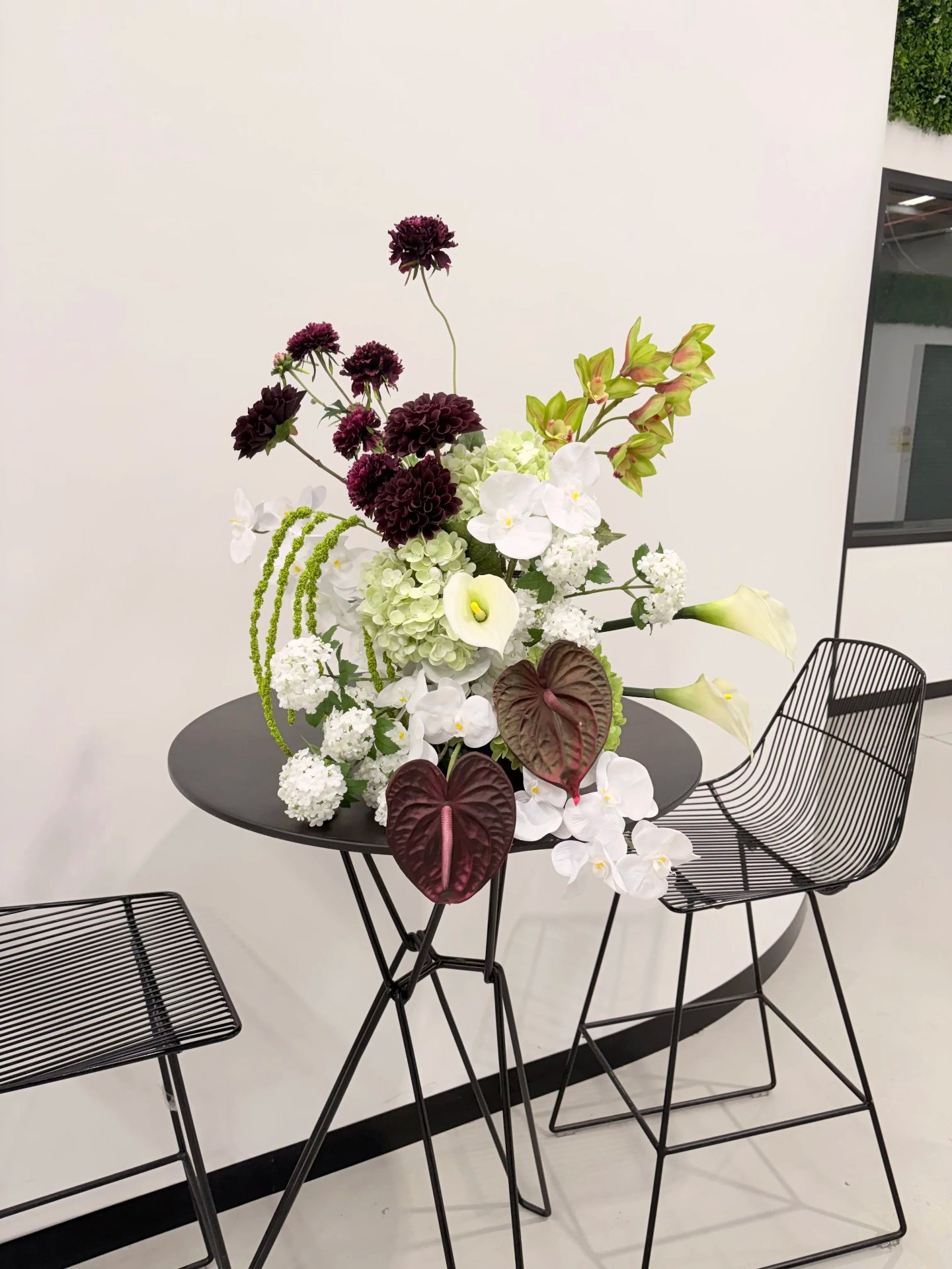 Sculptural artificial flower arrangement in green, burgundy and white colours black bar table with black wire chairs