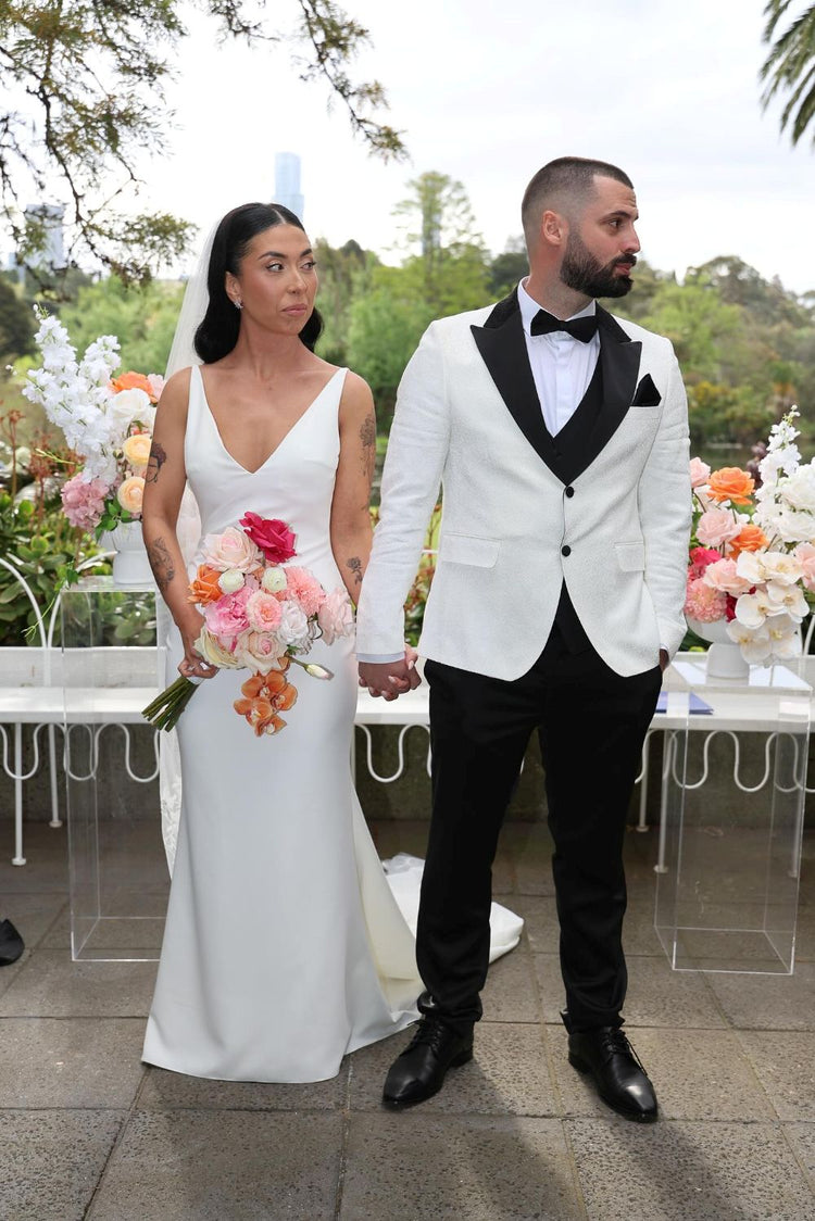 Bride and groom in wedding attire standing together outdoors with floral arrangements.