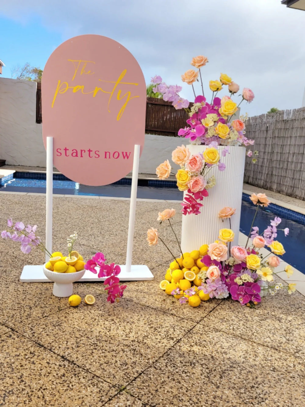 Pink welcome sign with a white stand surrounded by florals and lemons with a pool in the background