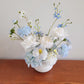 Light blue and white flower arrangement on a wooden bench against a white wall