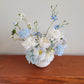 Light blue and white artificial flower arrangement on a wooden bench against a white wall