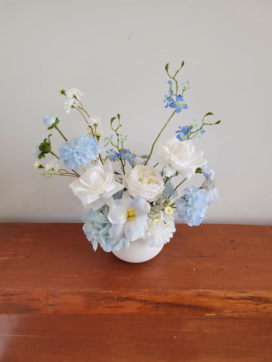 Light blue and white artificial flower arrangement on a wooden bench against a white wall
