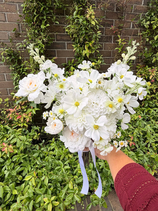 Faux wedding bouquet of white flowers held by a person against a brick wall and greenery background
