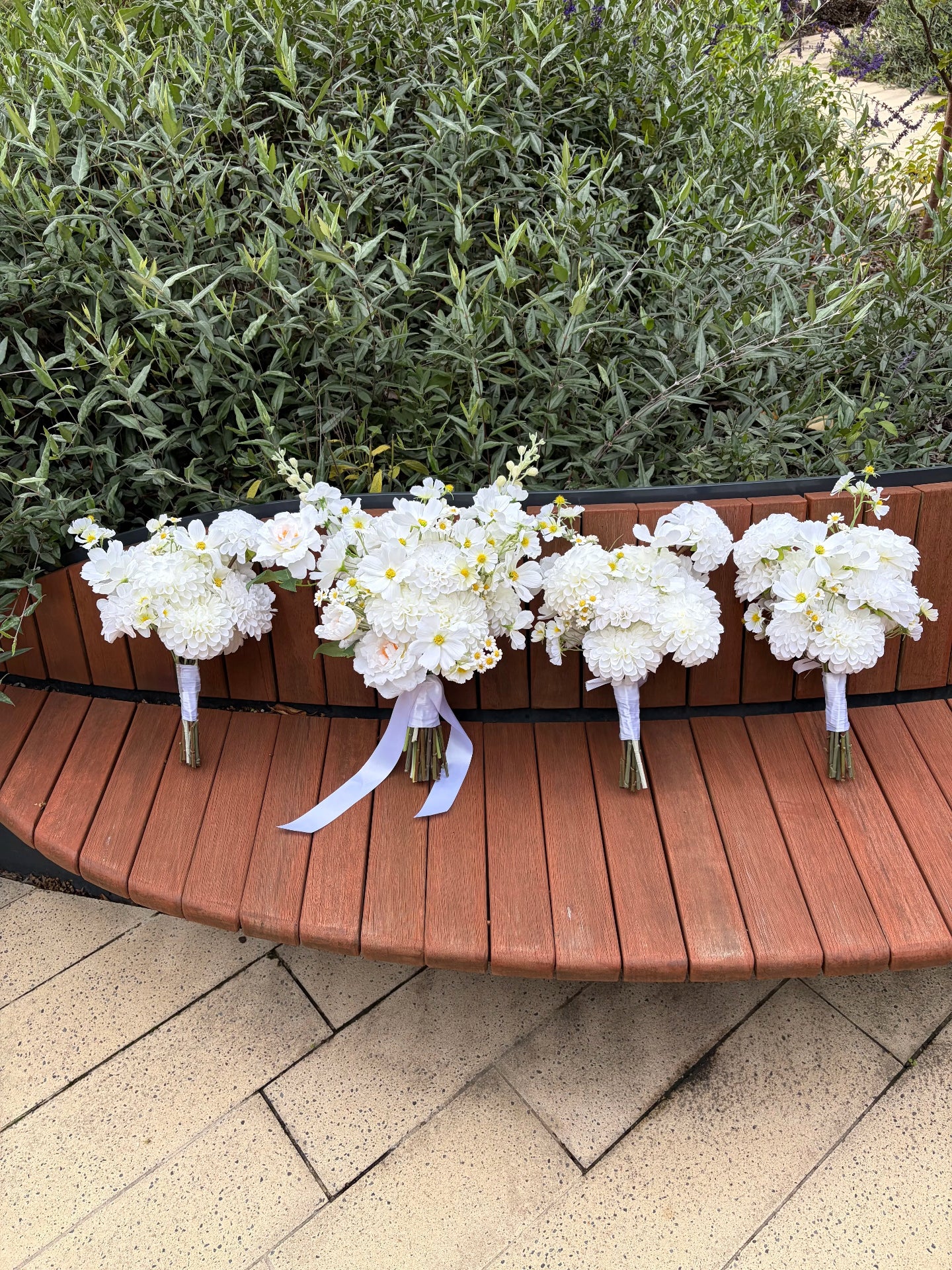 Four white artificial floral bouquets on a wooden bench with greenery in the background