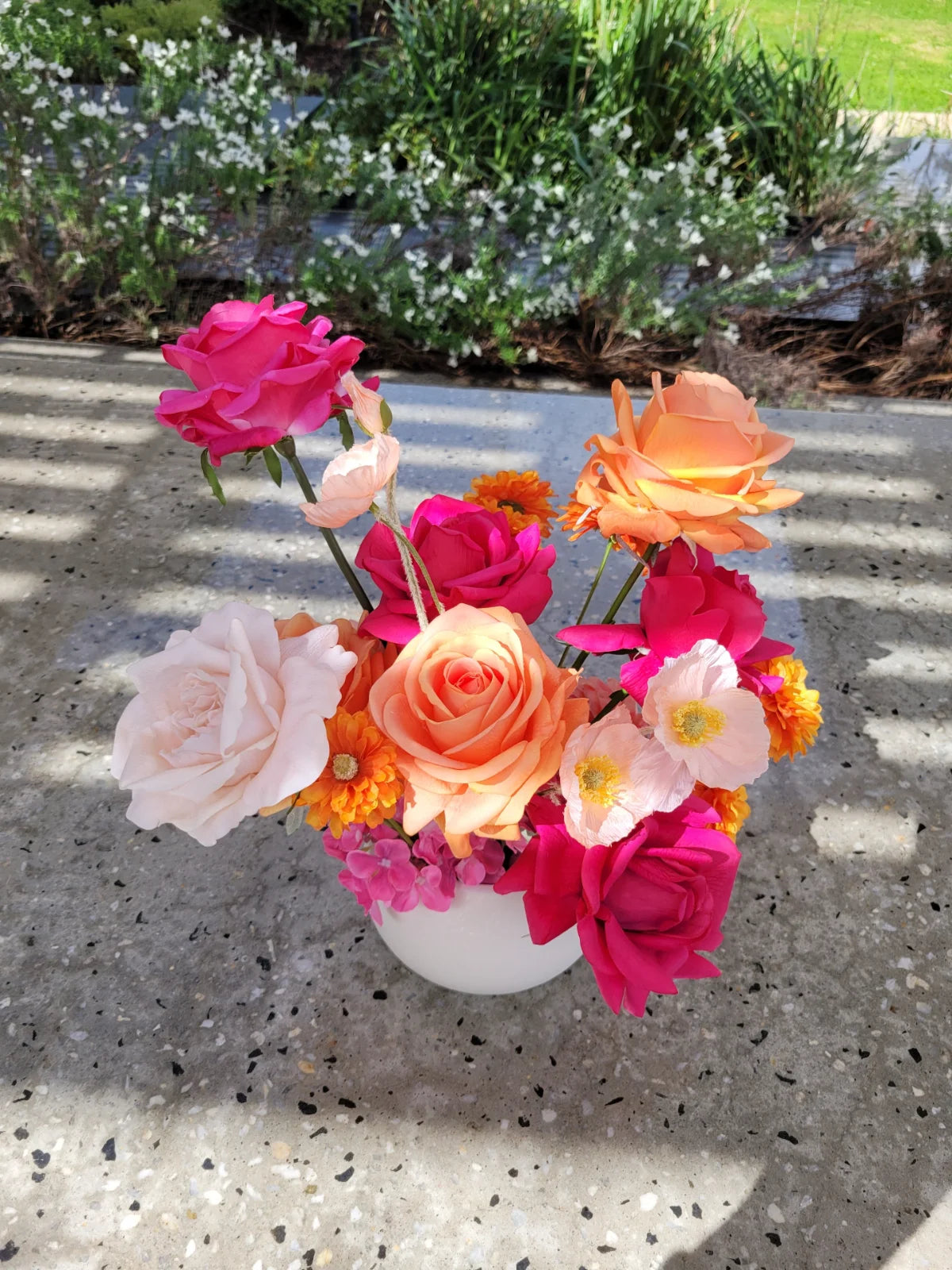 Colourful flower arrangement in a white vase on a concrete surface with greenery in the background.