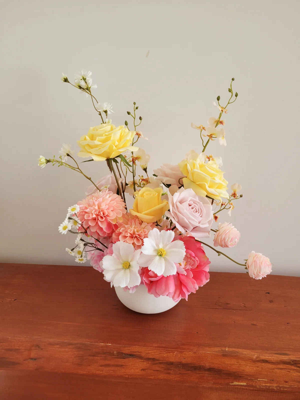 Floral arrangement with yellow, pink, and white flowers in a white vase on a wooden surface.