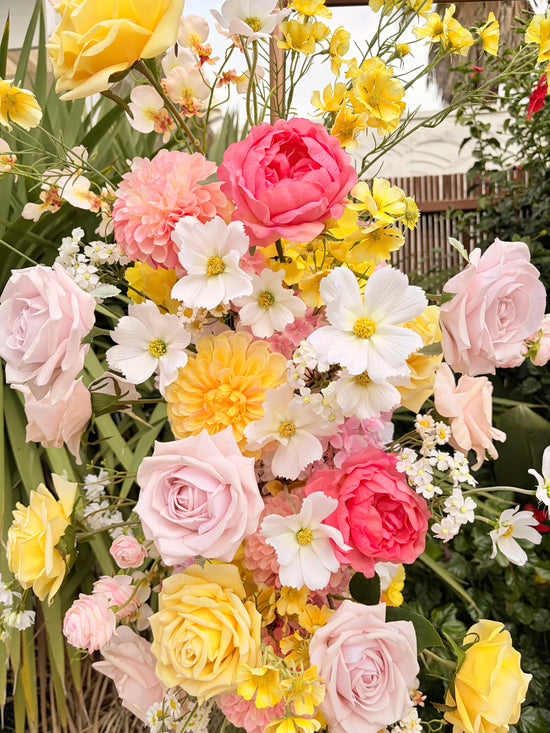 Close up of a wedding arbour artificial flower arrangement in pink and yellow with white cosmos in a garden setting